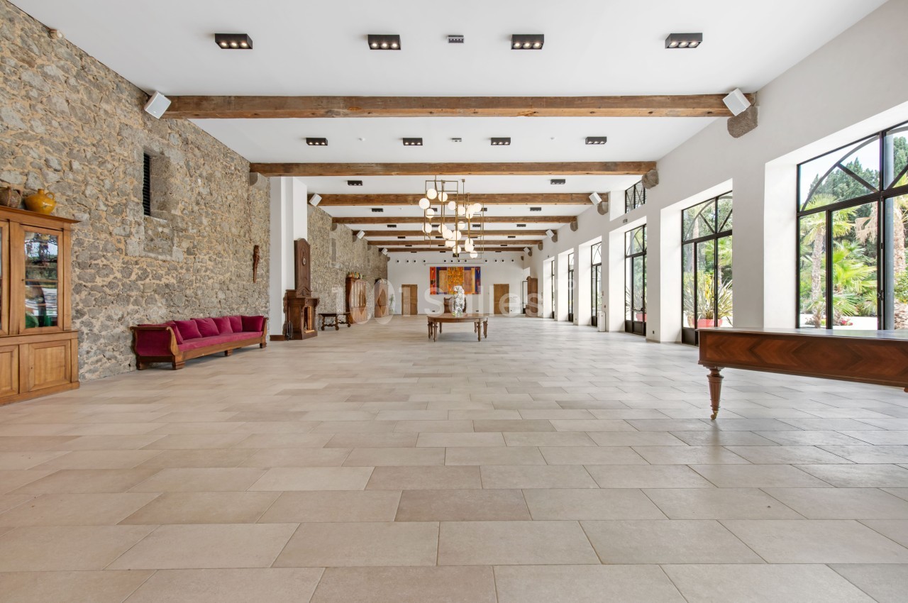 Grande salle lumineuse avec murs en pierre, poutres apparentes, mobilier en bois et larges baies vitrées donnant sur l…