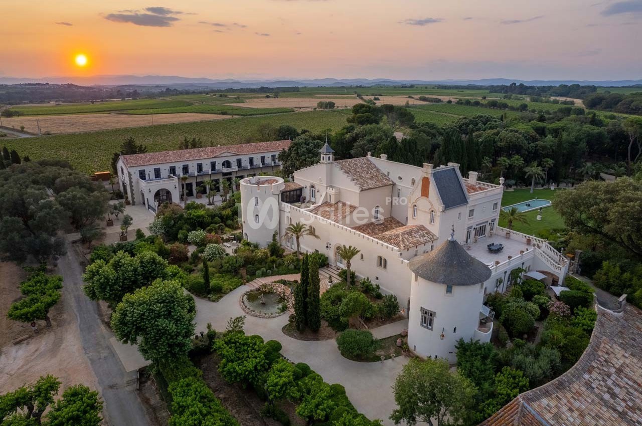 Vue aérienne d'un domaine avec un château entouré de jardins, de végétation et de champs au coucher du soleil.