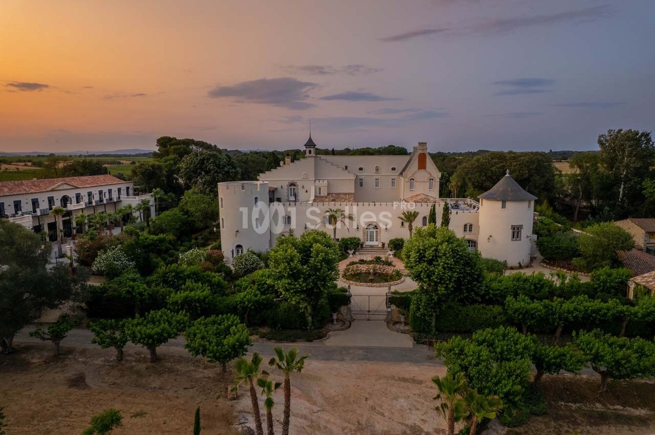 Vue aérienne d'un château entouré de jardins verdoyants au coucher du soleil, avec des bâtiments annexes à proximité.