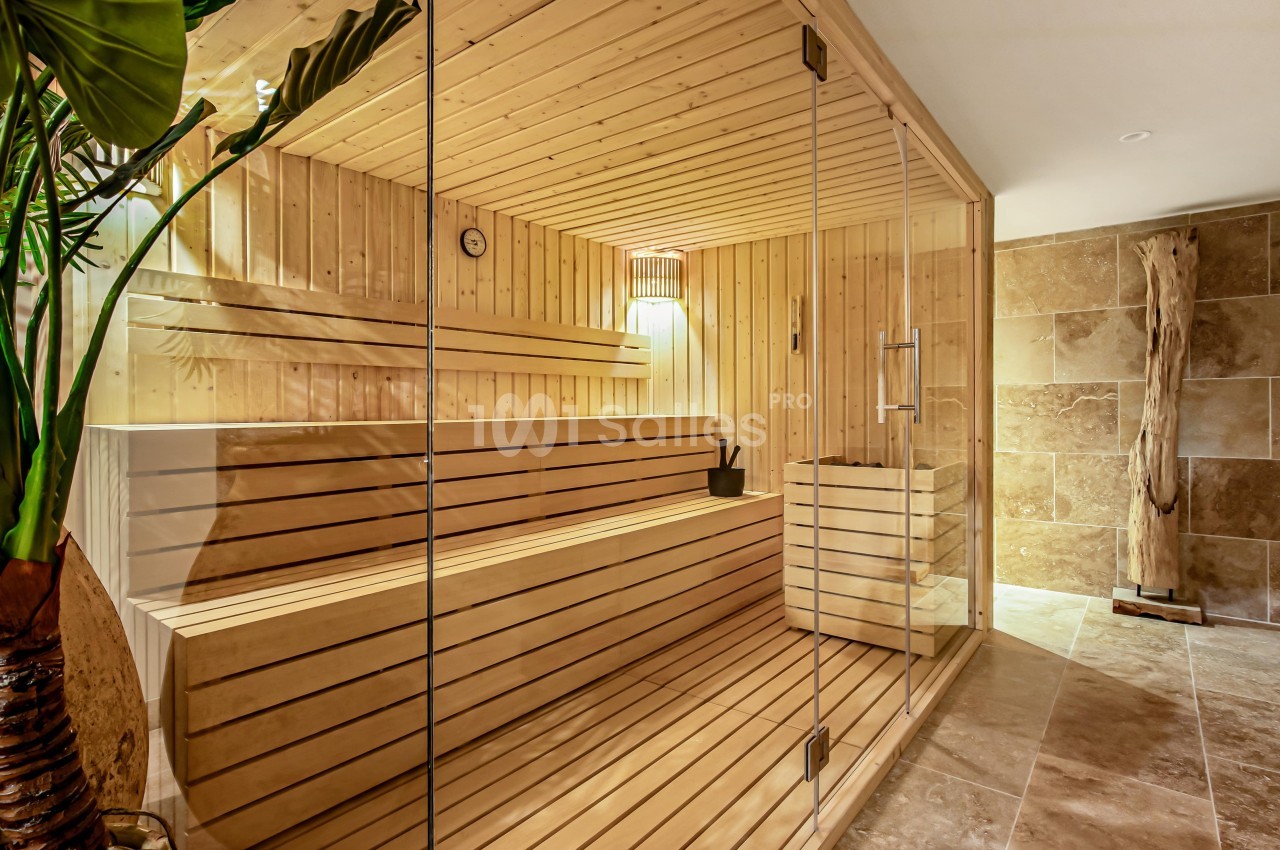 Sauna en bois clair avec bancs, seau et louche, éclairé par une lampe, entouré de murs en pierre beige.
