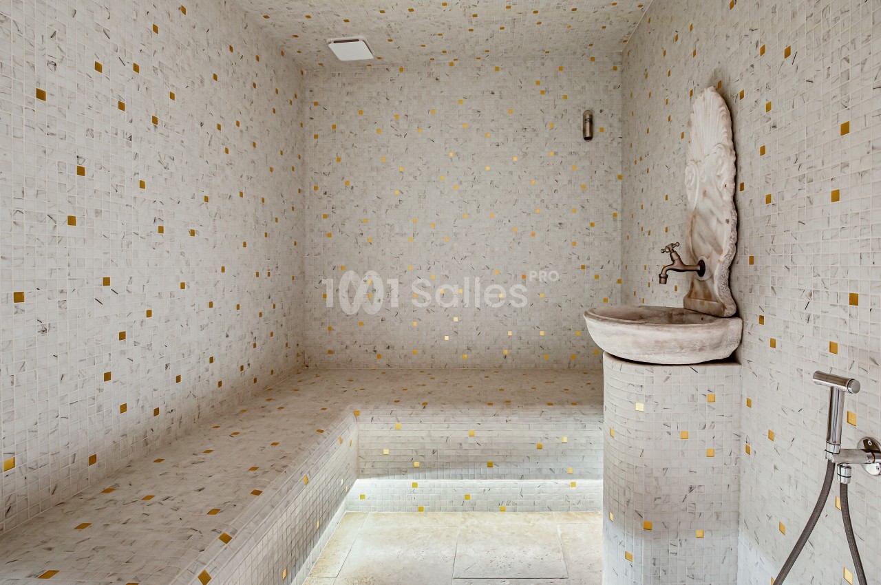 Hammam carrelé de mosaïques blanches et dorées avec une banquette intégrée et une fontaine murale sculptée.