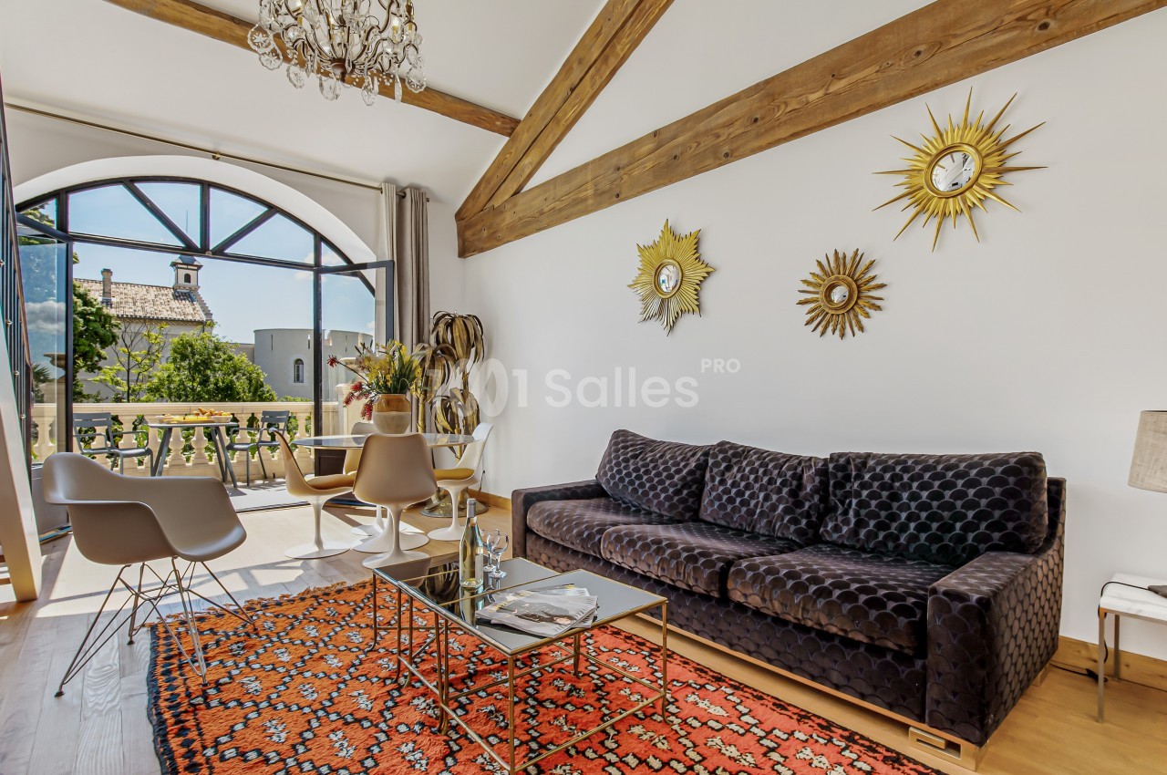 Salon lumineux avec canapé, table basse, tapis coloré, chaises modernes, grande baie vitrée et vue sur un clocher.