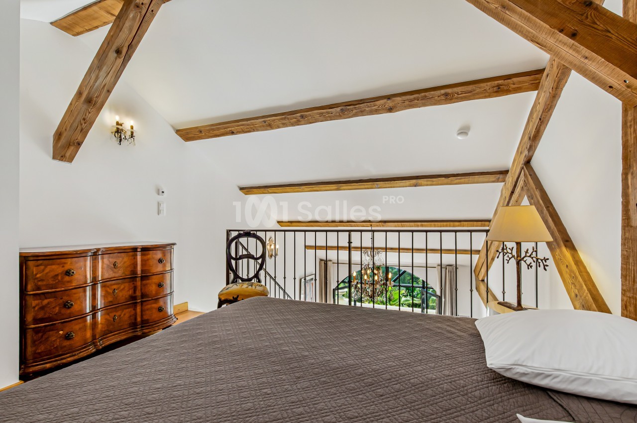 Chambre mansardée avec poutres apparentes, lit double, commode en bois et vue sur une fenêtre en arche.