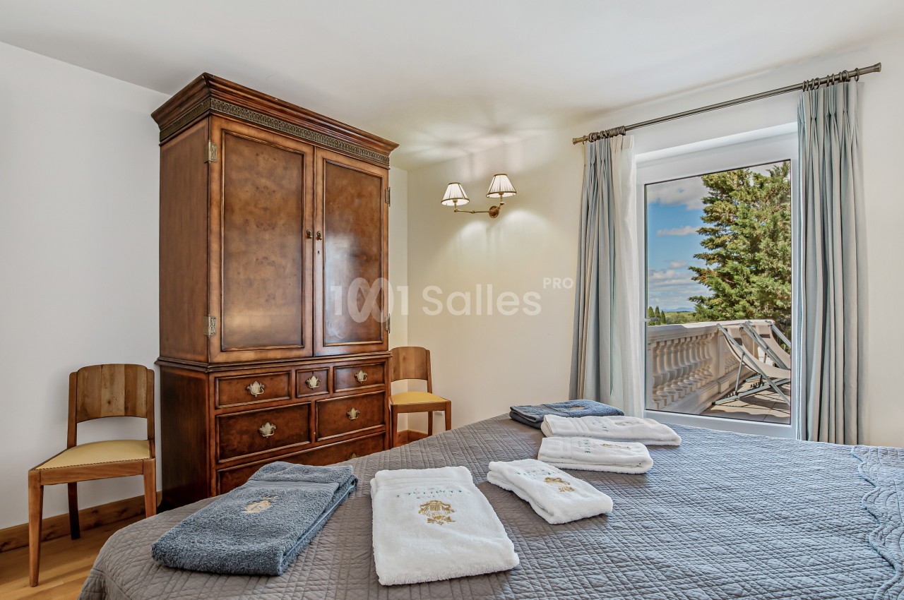 Chambre lumineuse avec armoire en bois, lit couvert de serviettes pliées et accès à un balcon avec vue sur des arbres.