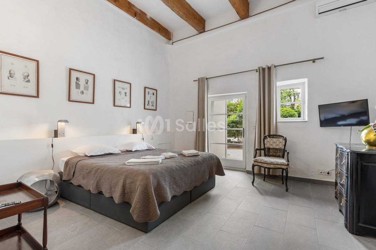 Chambre lumineuse avec lit double, murs blancs, poutres apparentes, fenêtres donnant sur un jardin et mobilier classique.