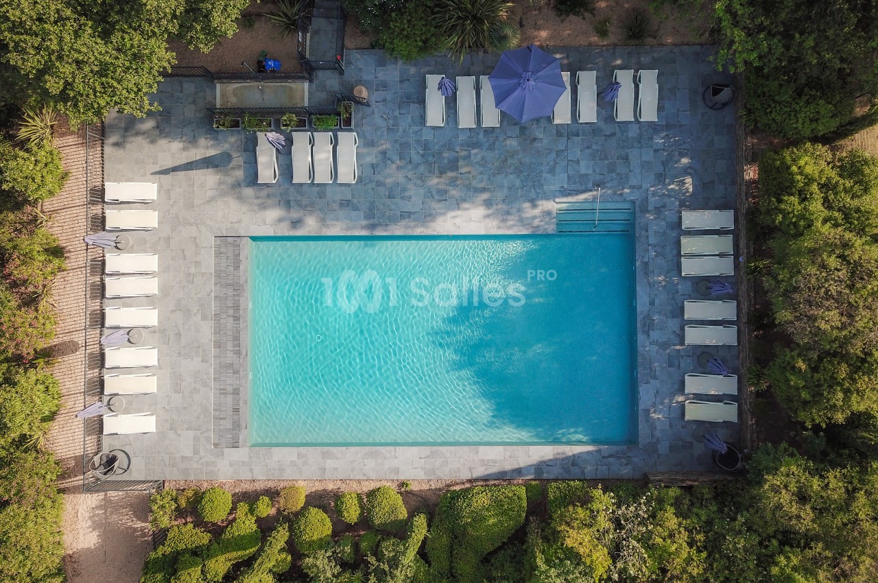 Vue aérienne d'une piscine rectangulaire entourée de chaises longues, parasols et végétation.