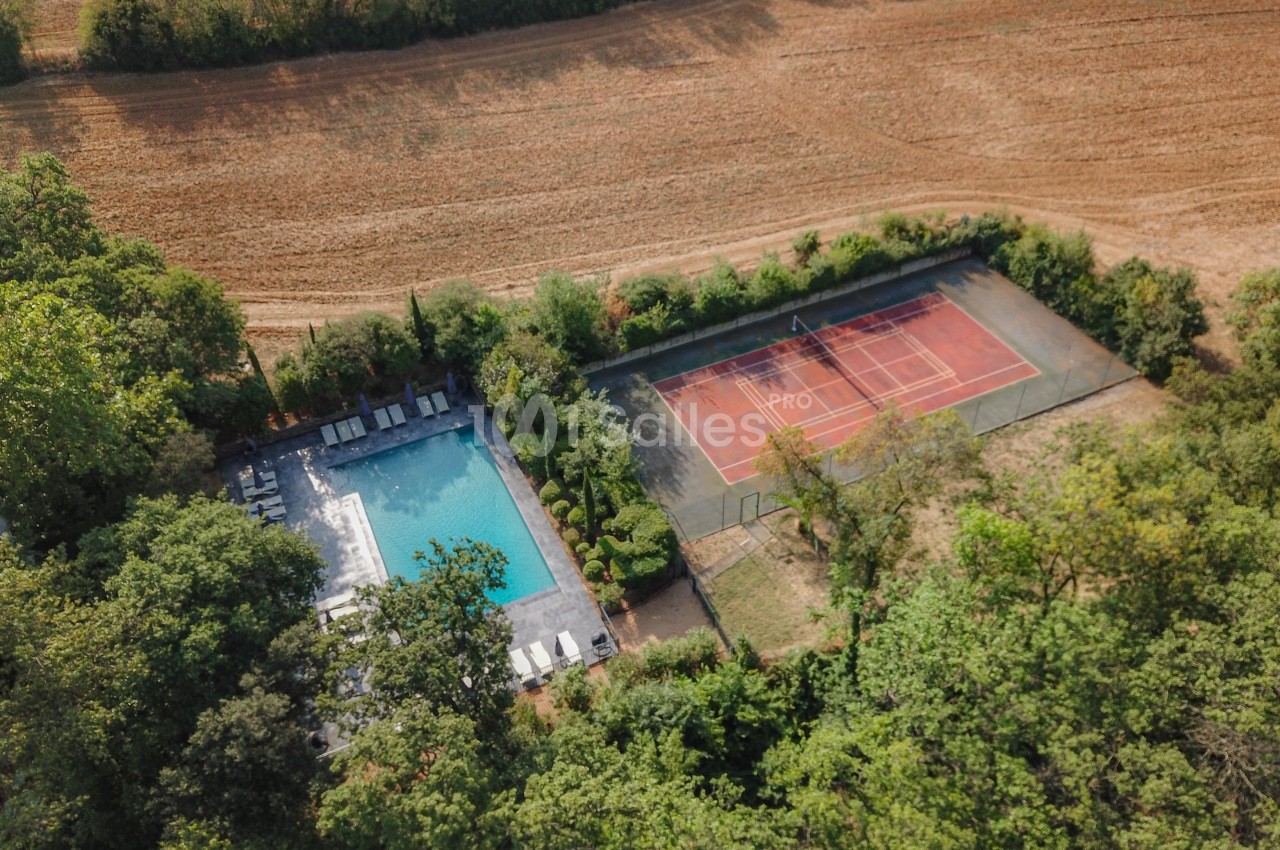 Vue aérienne d'une piscine entourée de chaises longues et d'un terrain de tennis au milieu d'un paysage rural.