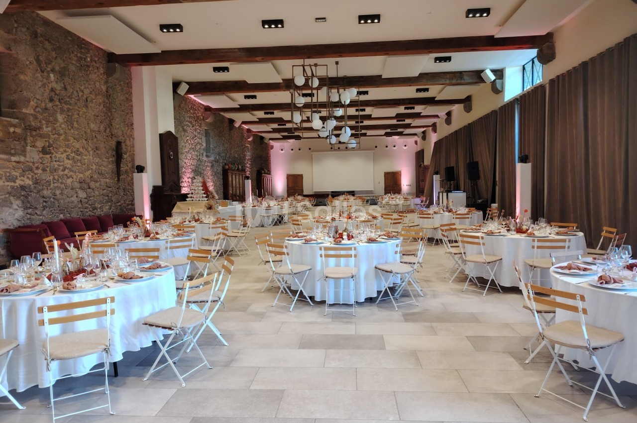 Salle de réception décorée avec des tables rondes dressées, chaises en bois et éclairage chaleureux.