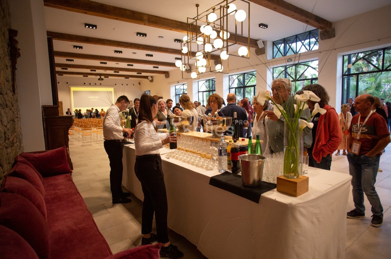 Des personnes se servent à un buffet avec boissons et verres, dans une salle lumineuse avec des tables et des lustres…