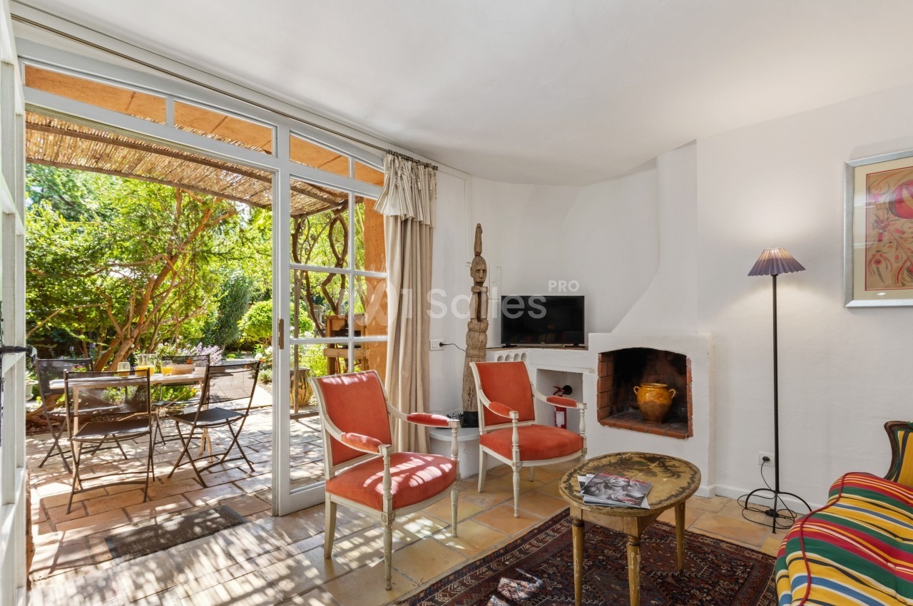 Salon lumineux avec cheminée, deux fauteuils, table basse et vue sur une terrasse ombragée avec table et chaises.