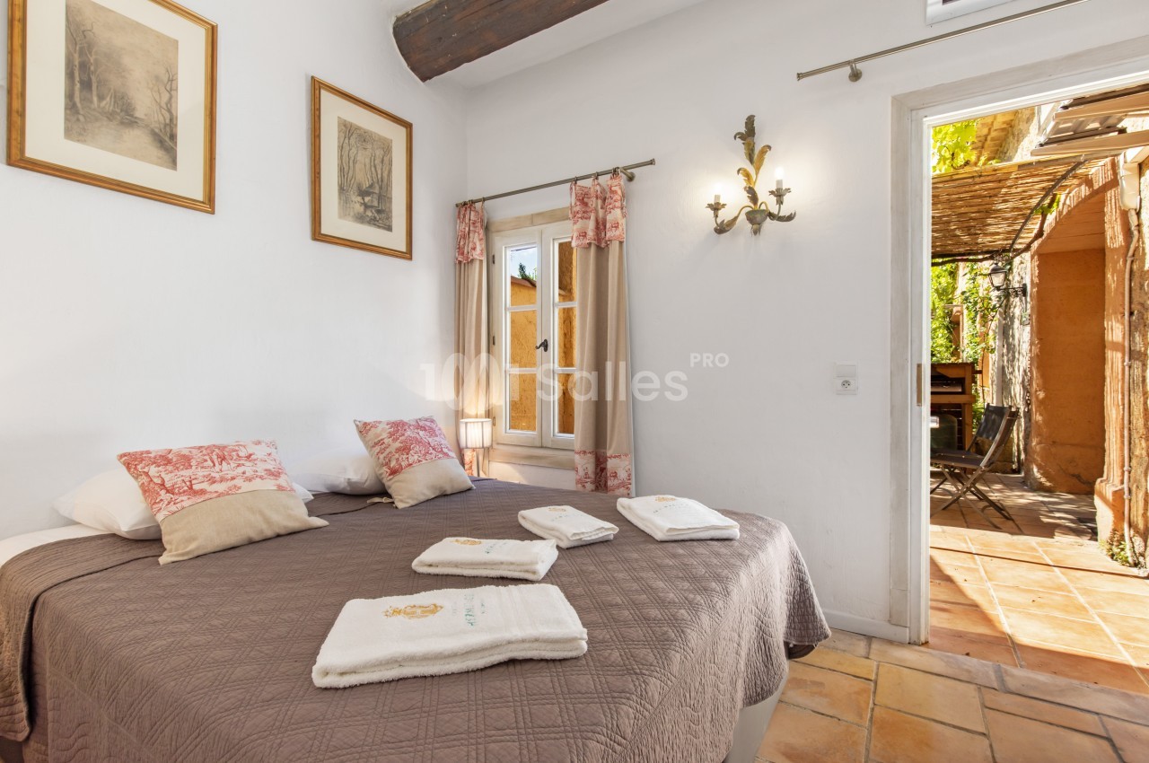 Chambre lumineuse avec lit double, décor rustique, poutre apparente, et accès à une terrasse ensoleillée.