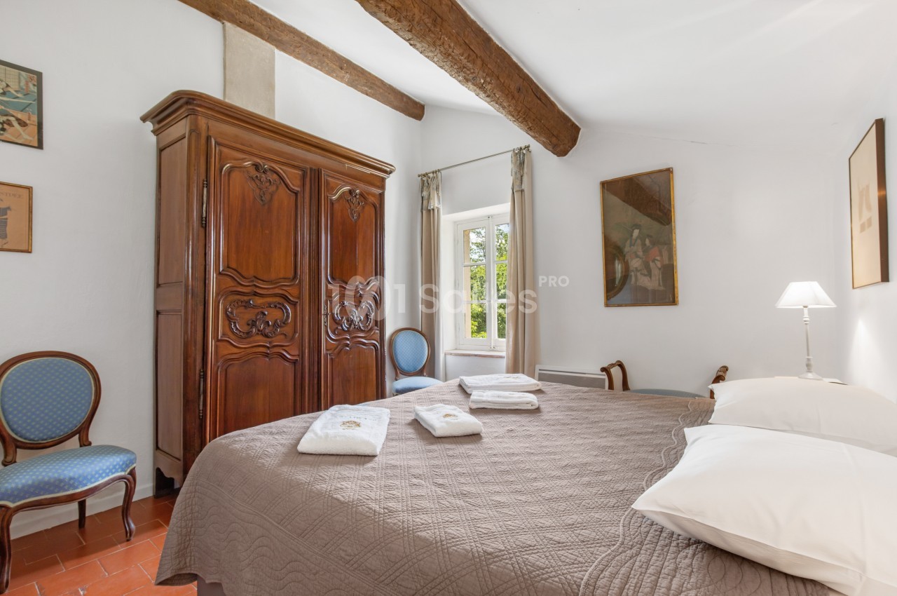 Chambre lumineuse avec lit double, armoire en bois sculpté, chaises bleues et fenêtre donnant sur un jardin.
