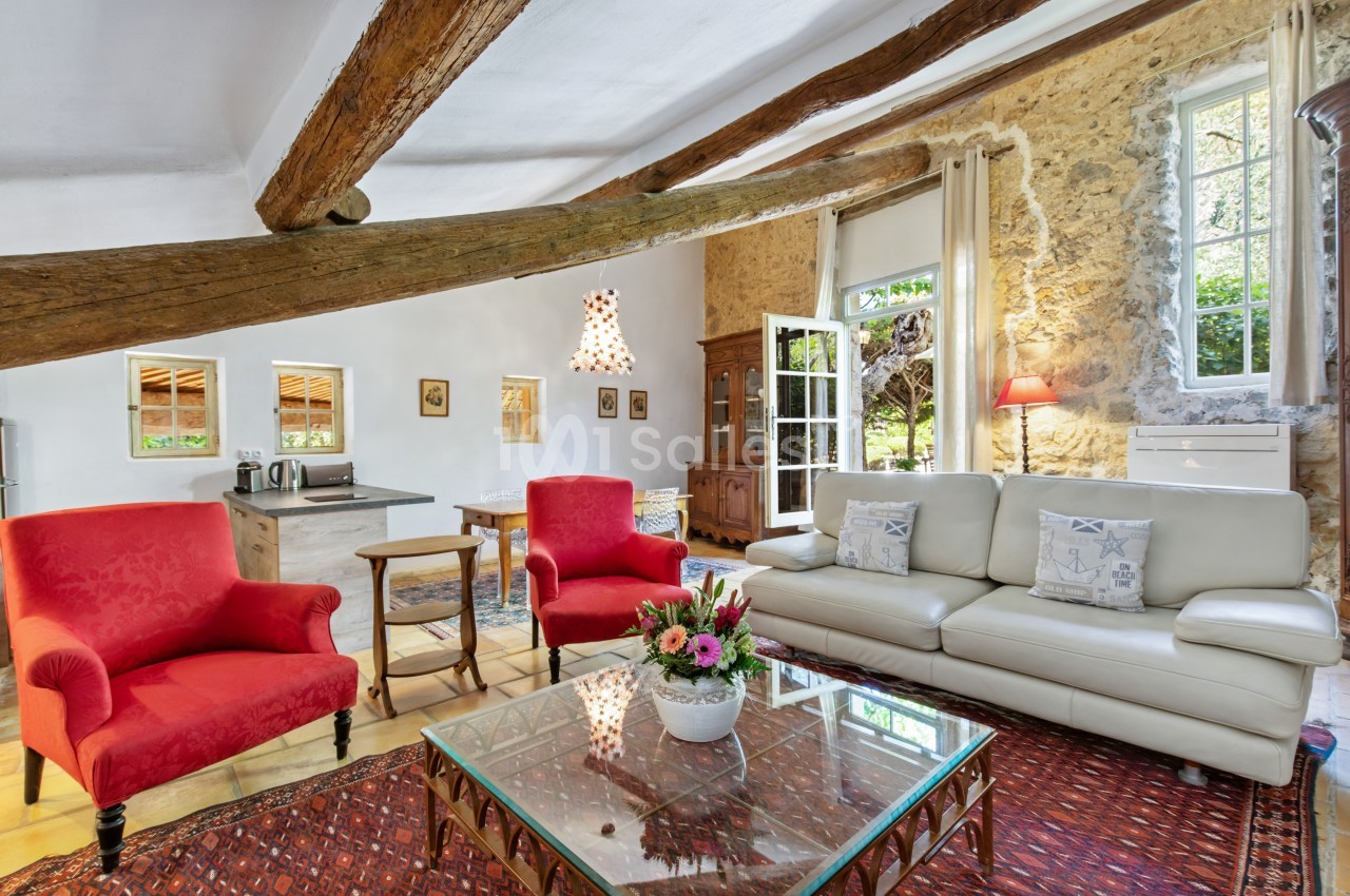 Salon lumineux avec poutres apparentes, canapé blanc, fauteuils rouges, table basse en verre et murs en pierre.