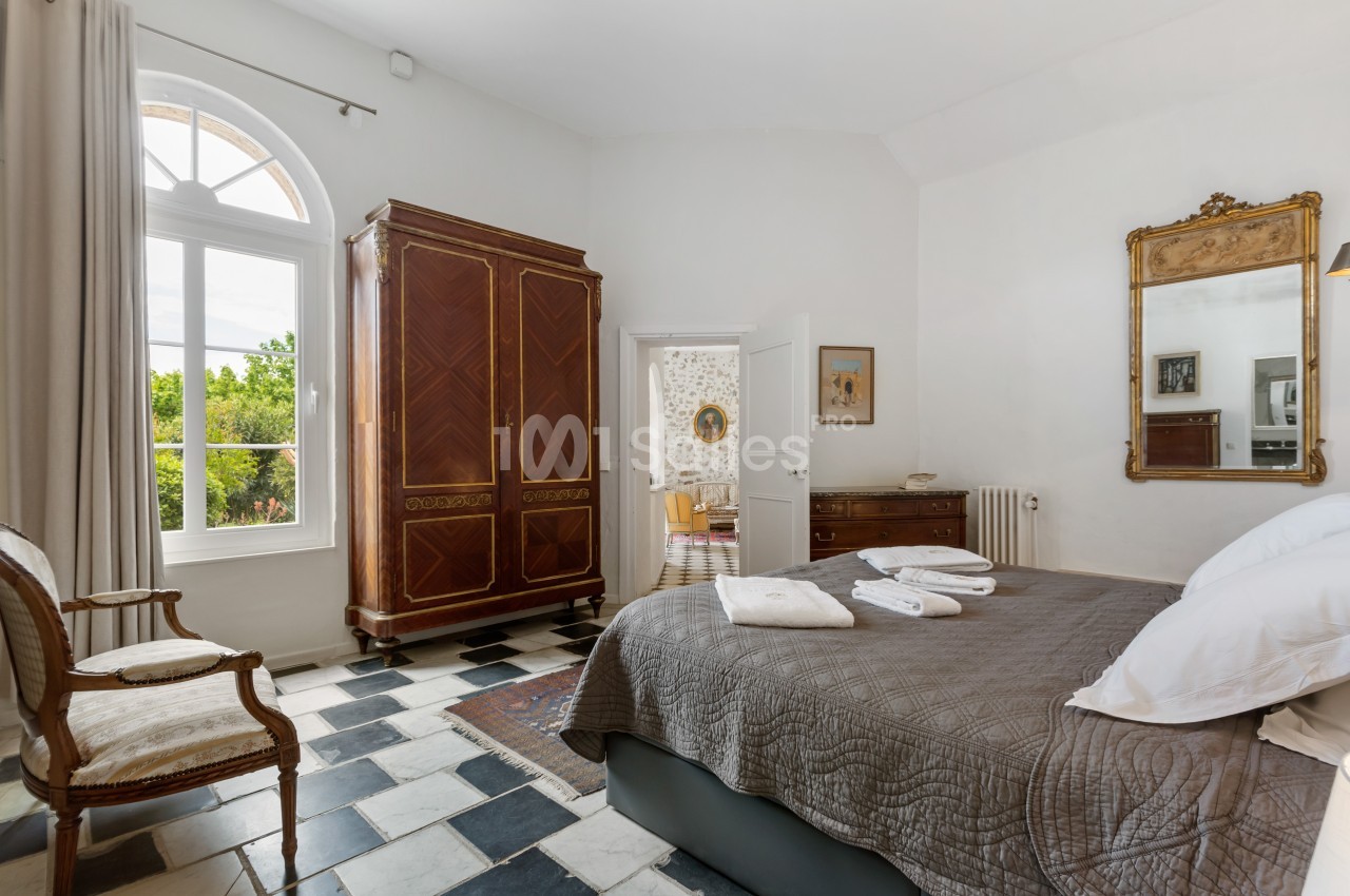 Chambre lumineuse avec lit double, armoire en bois, miroir doré, fenêtre cintrée et sol en carreaux noirs et blancs.