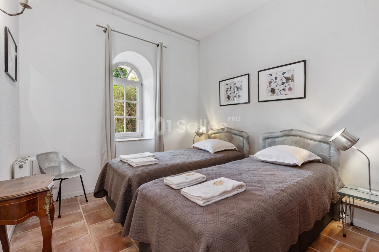 Chambre lumineuse avec deux lits simples, murs blancs, sol en terre cuite et fenêtre donnant sur un jardin.