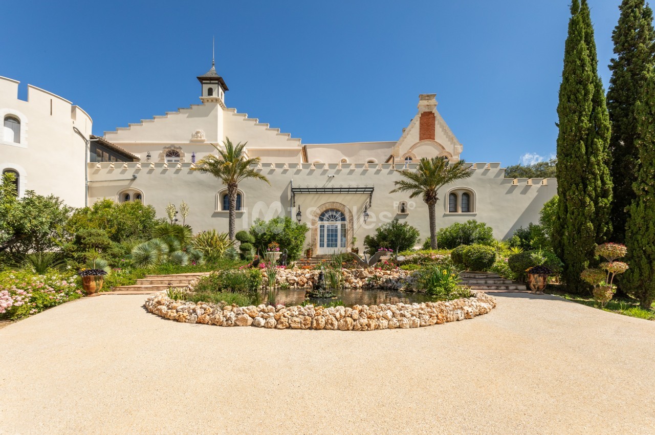Façade d'un bâtiment historique entouré de jardins fleuris, avec un ciel bleu dégagé en arrière-plan.