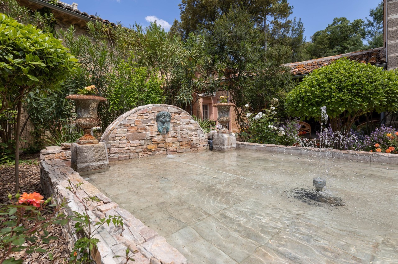 Fontaine en pierre dans un jardin méditerranéen entouré de végétation, avec une maison en arrière-plan.