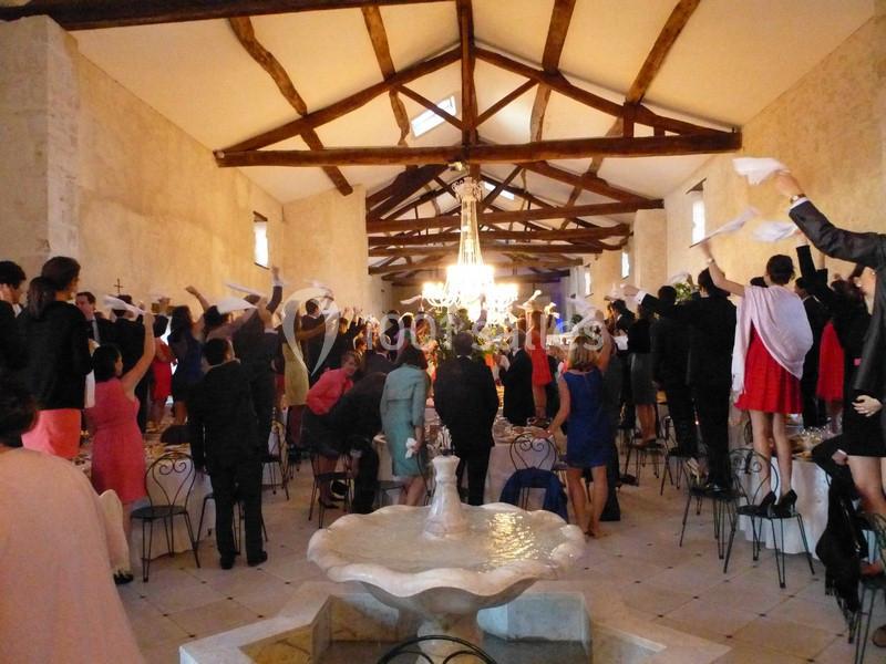Des invités debout agitent des serviettes blanches dans une salle de réception avec poutres apparentes et lustre central.