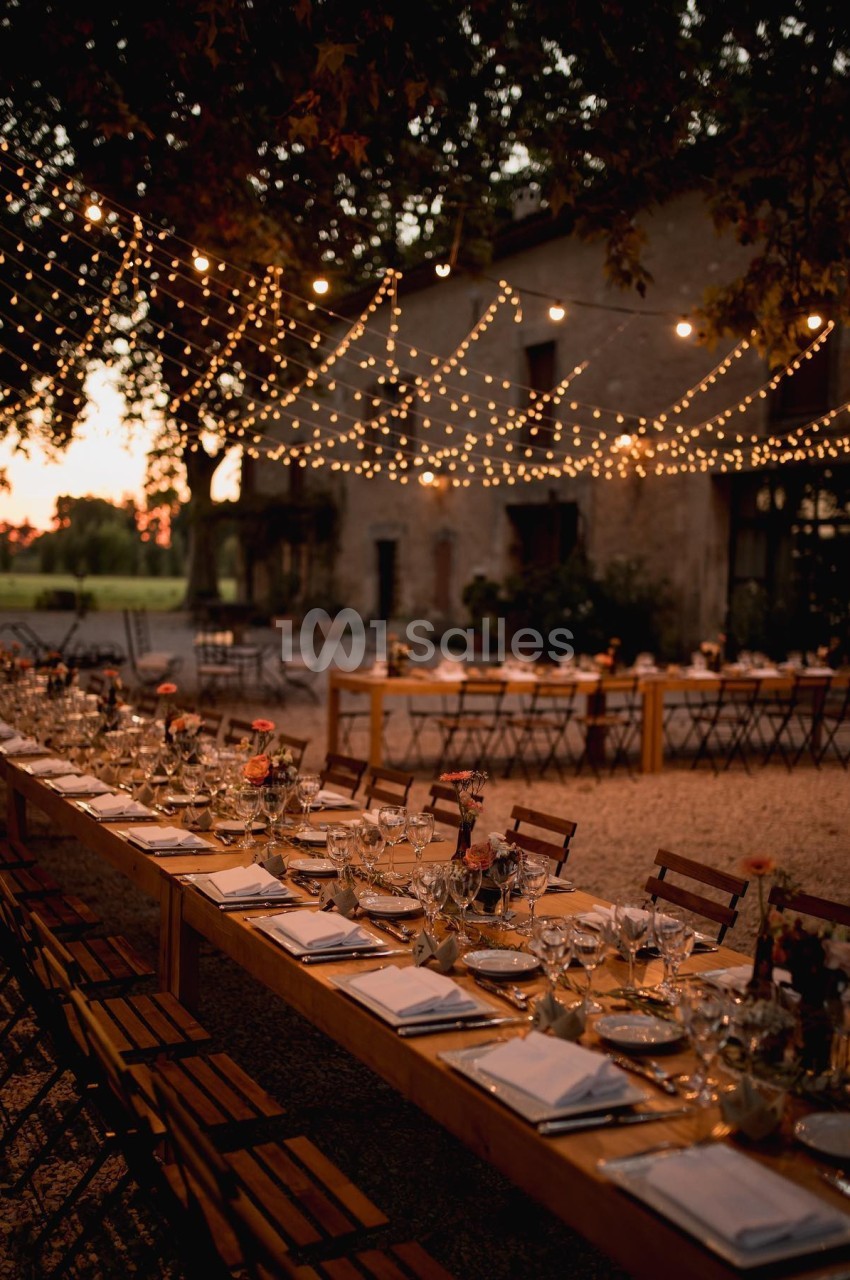 Tables en bois dressées pour un dîner en extérieur, éclairées par des guirlandes lumineuses au crépuscule.