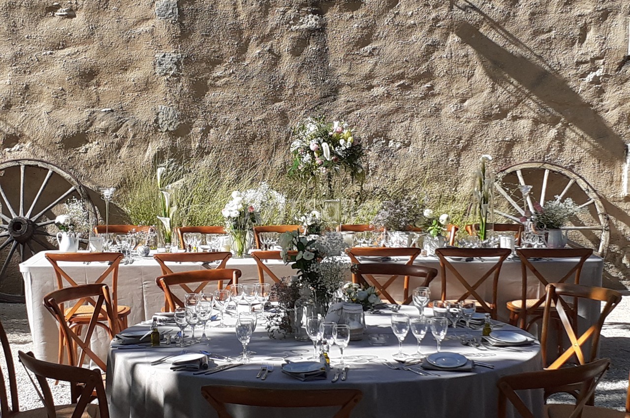 Tables dressées pour un repas en extérieur, avec nappes blanches, chaises en bois et décor floral devant un mur en pierre.