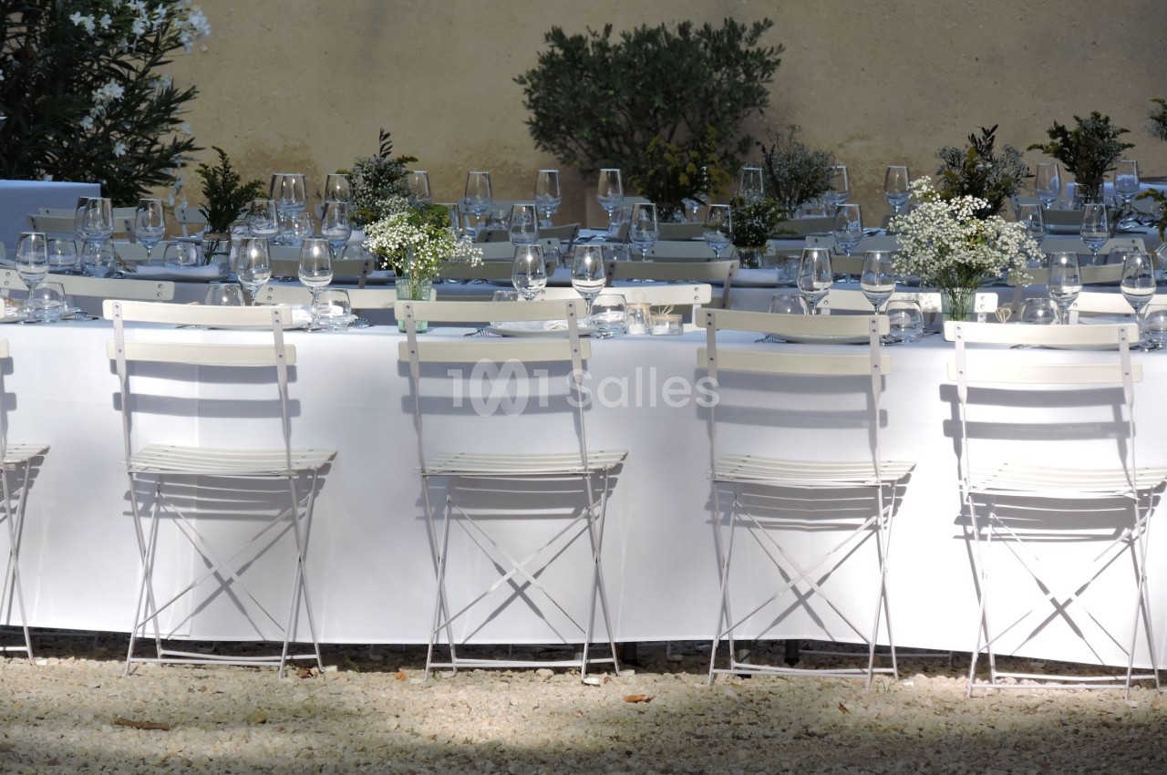 Table dressée en extérieur avec nappes blanches, chaises pliantes, verres, couverts et décorations florales.