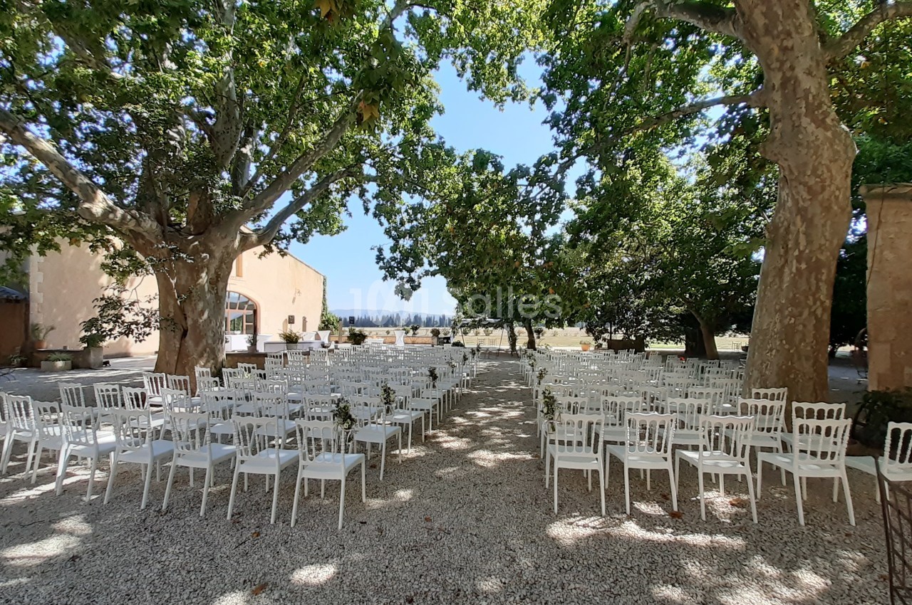 Chaises blanches disposées en rangées sous des arbres dans une cour extérieure ensoleillée, préparée pour un événement.