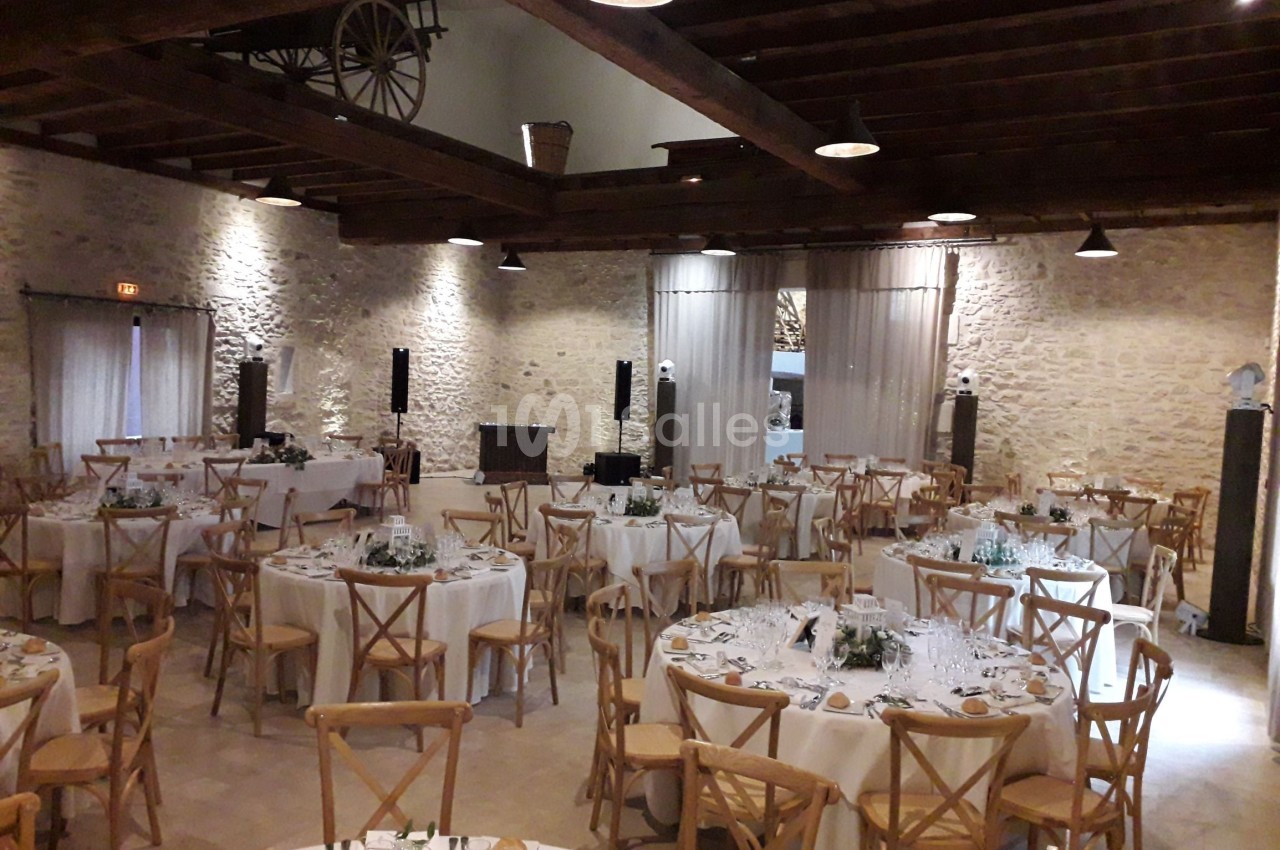 Salle de réception avec des tables rondes dressées, chaises en bois et murs en pierre apparente.