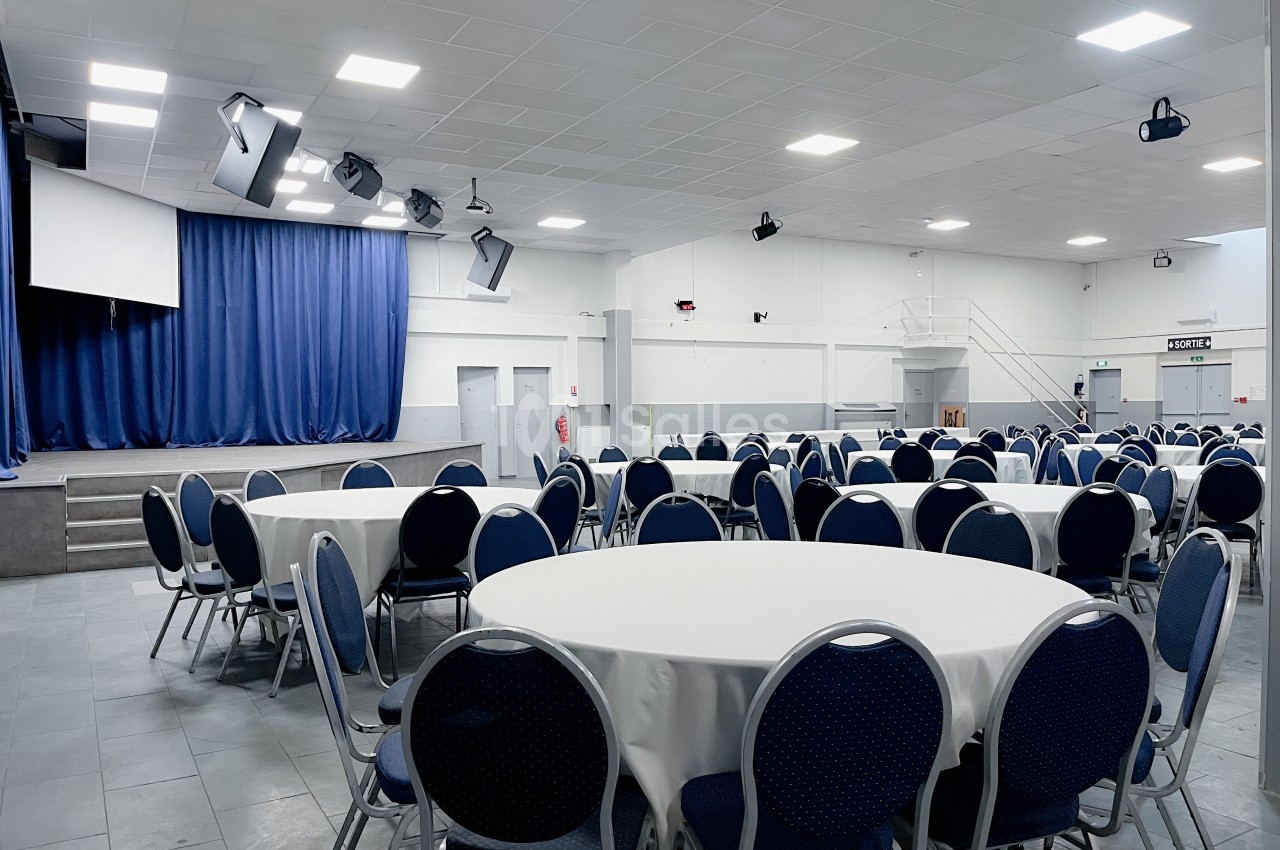 Salle de réception avec des tables rondes dressées de nappes blanches, une scène avec rideaux bleus en arrière-plan.