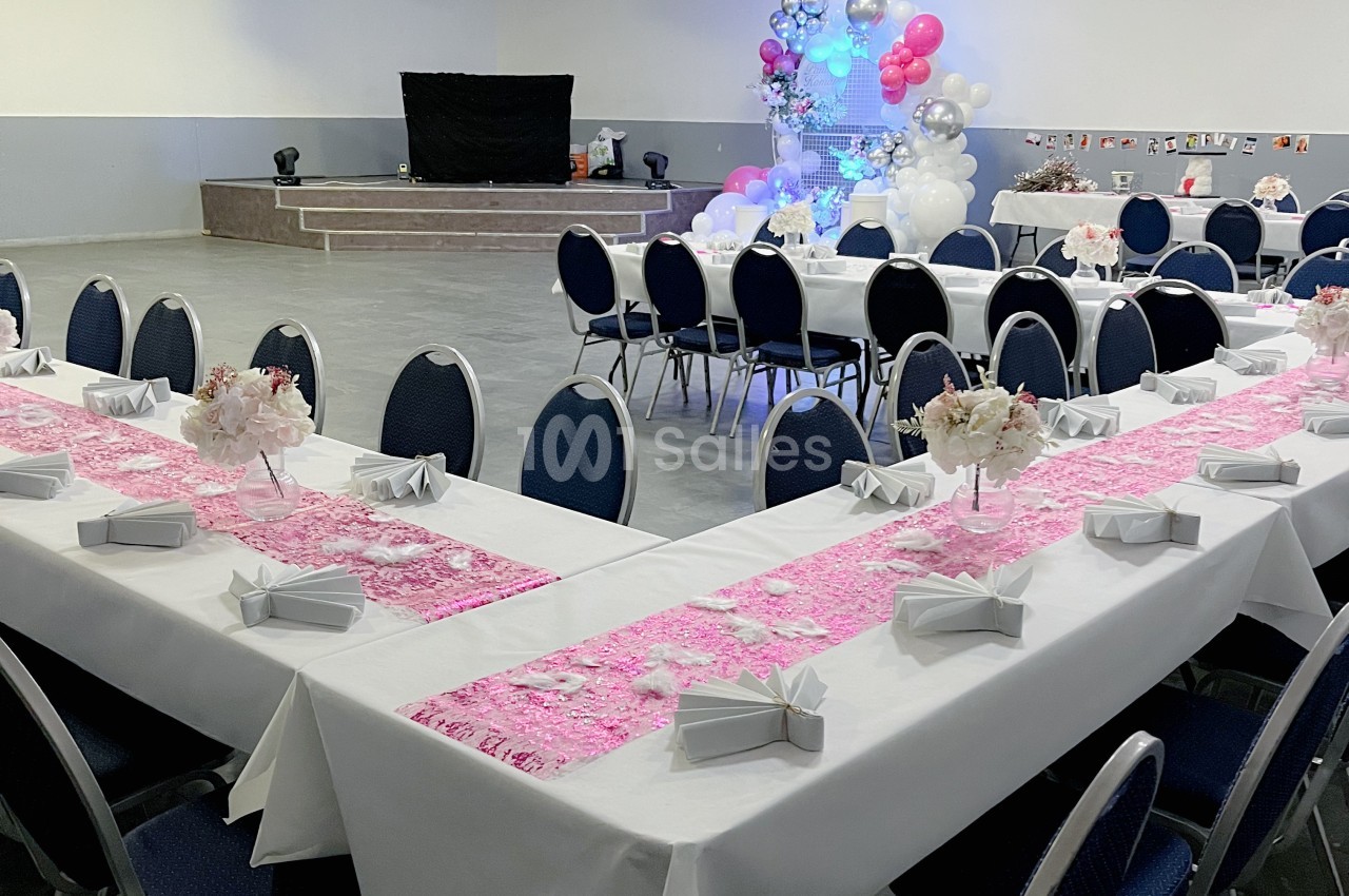 Salle de réception décorée avec des tables en U, nappes blanches, chemins de table roses et ballons en arrière-plan.