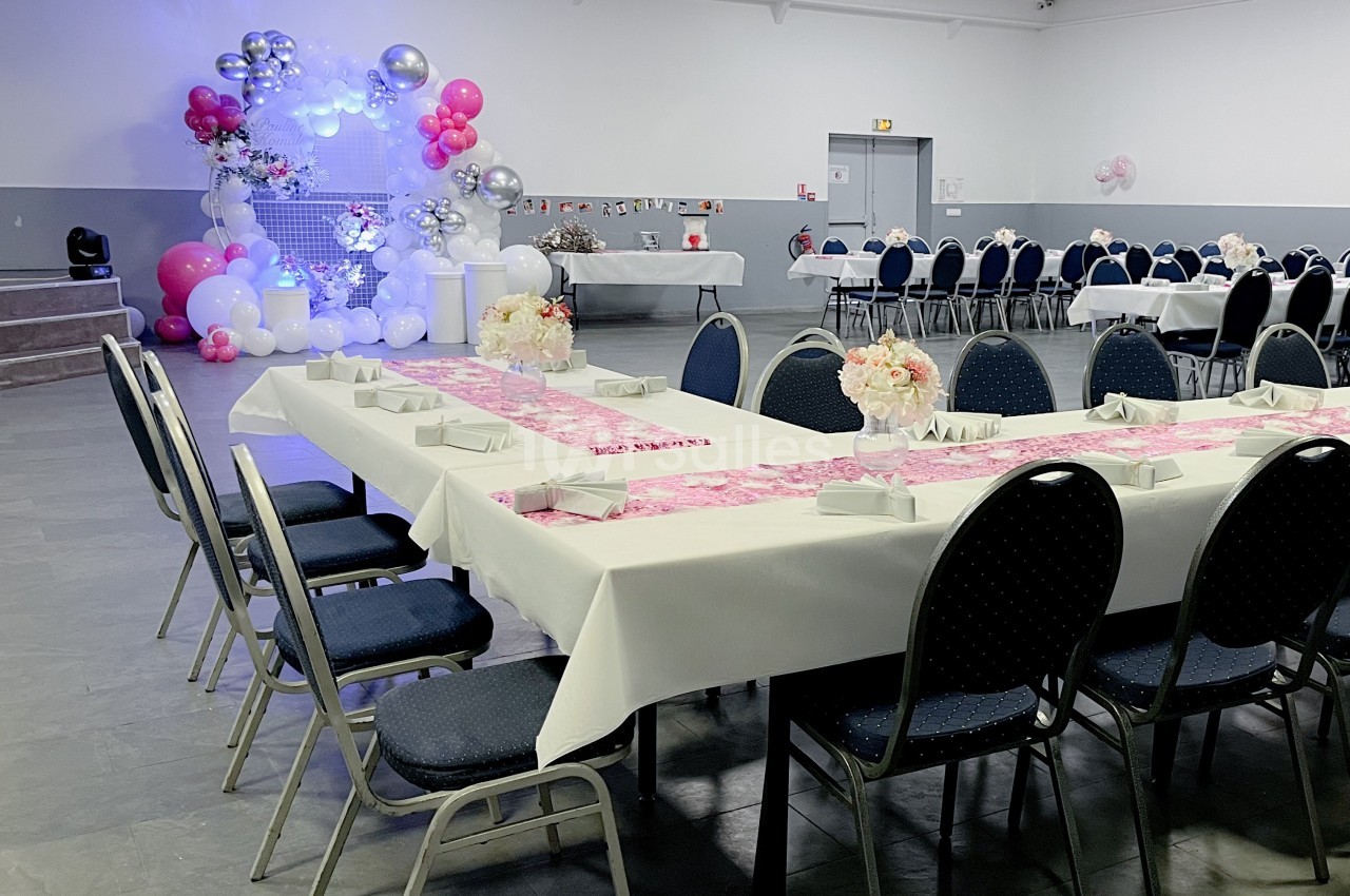 Salle décorée pour un événement avec tables dressées, chaises alignées et arche de ballons roses et argentés en arrière-plan.