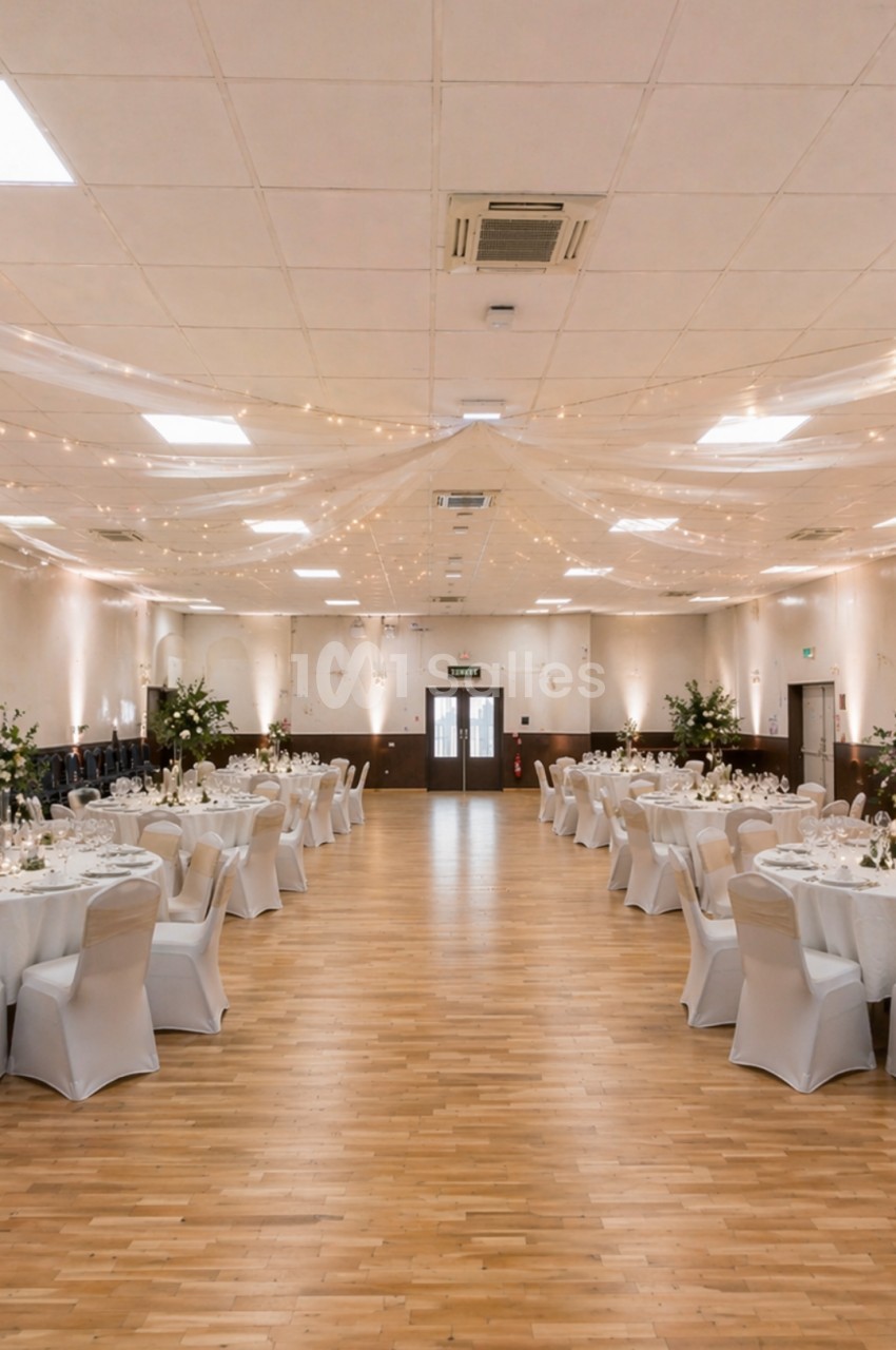 Salle de réception décorée avec des tables rondes dressées, chaises blanches et guirlandes lumineuses suspendues.