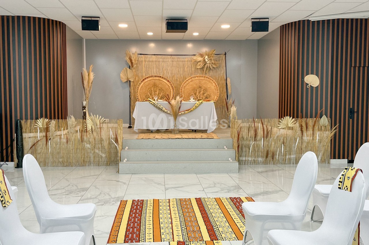 Salle décorée pour un événement avec des chaises blanches, tapis colorés et scène ornée d'éléments naturels.