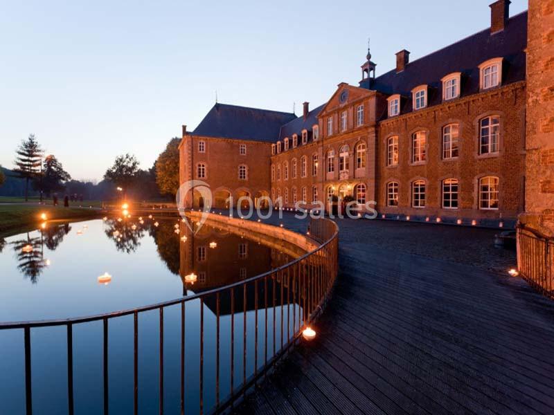 Façade d'un château en pierre éclairé au crépuscule, avec un étang et une passerelle en bois au premier plan.