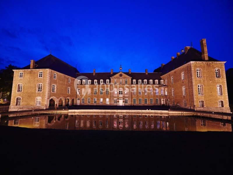 Façade d'un château éclairé la nuit, reflété dans un plan d'eau calme sous un ciel bleu profond.