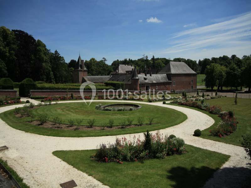 Jardin paysager avec parterres fleuris, allées gravillonnées et bâtiment en briques rouges sous un ciel dégagé.