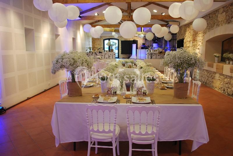 Salle décorée pour un événement avec tables dressées, chaises blanches, fleurs blanches et lanternes suspendues.