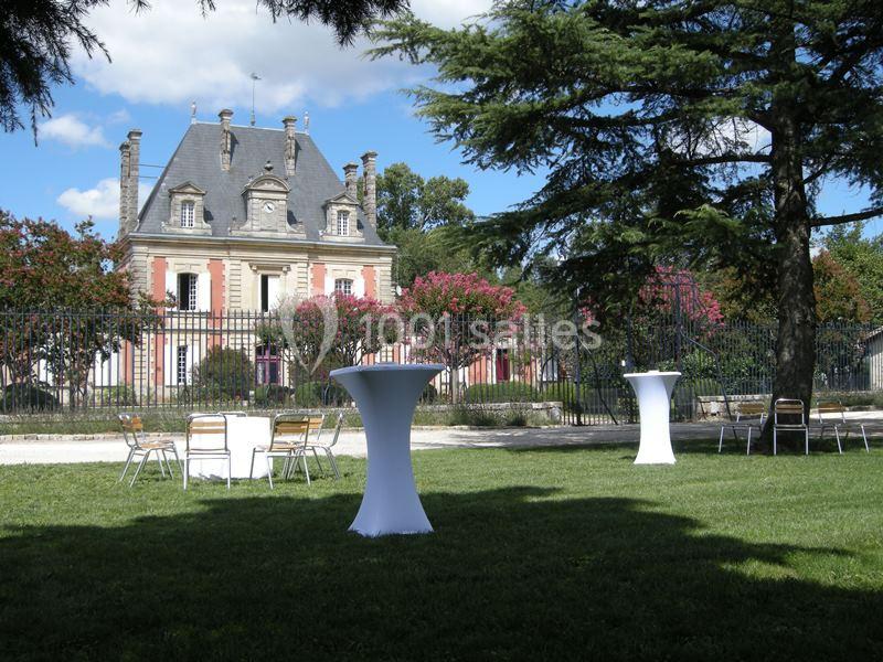 Jardin avec tables hautes et chaises disposées devant un bâtiment ancien entouré d'arbres et de grilles.