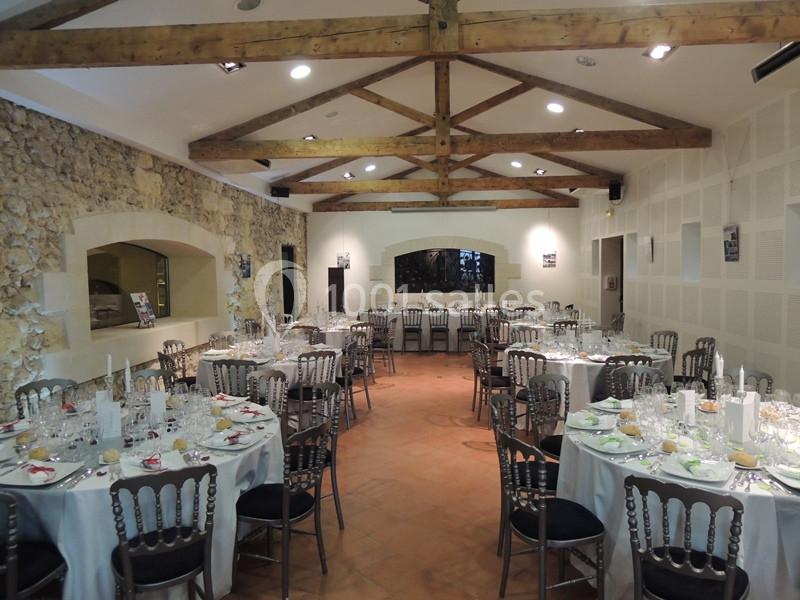 Salle de réception avec tables rondes dressées, chaises en métal et poutres apparentes au plafond.
