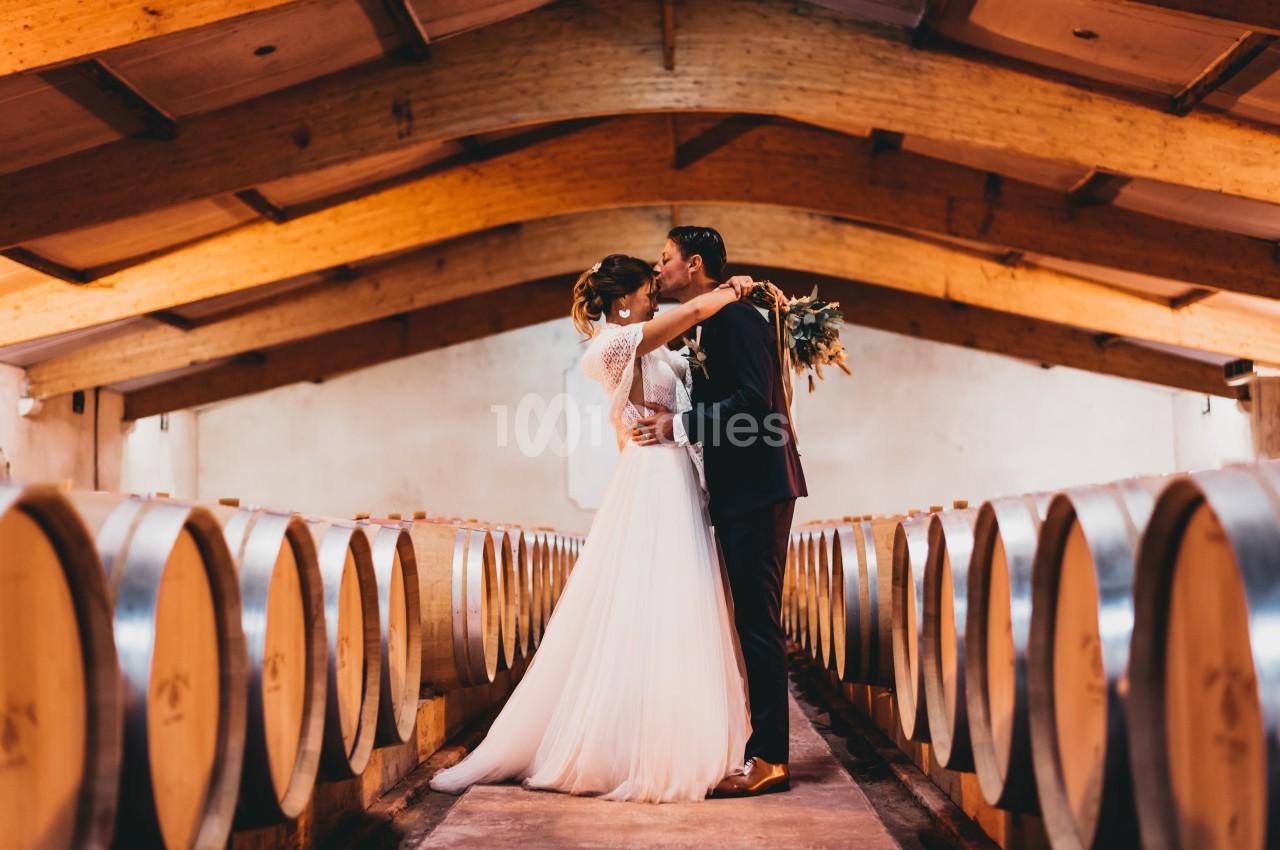 Un couple en tenue de mariage s'embrasse dans une cave à vin entourée de tonneaux en bois.