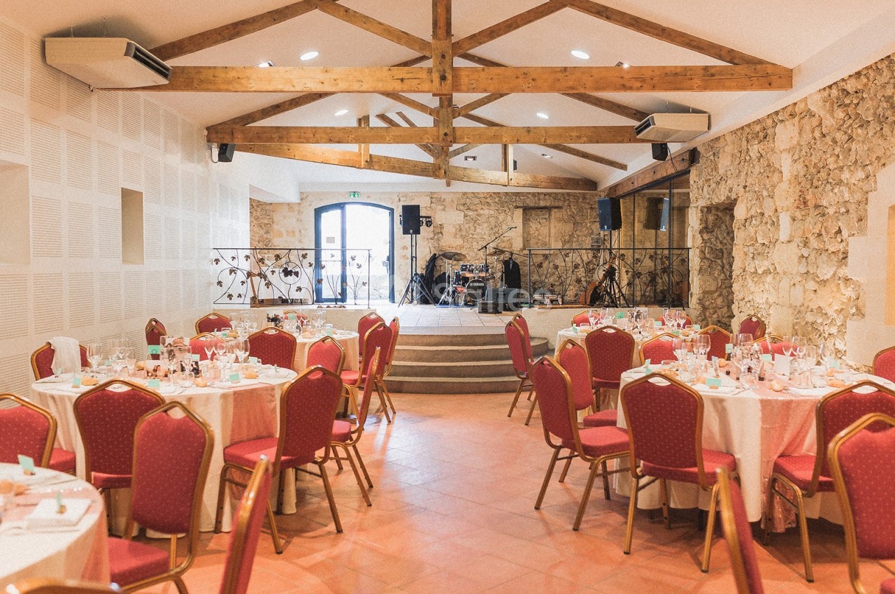 Salle de réception avec tables rondes dressées, chaises rouges et scène équipée de matériel audio.