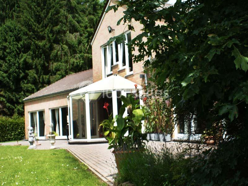 Maison en briques avec une véranda, entourée de végétation et d'arbres, vue depuis un jardin.