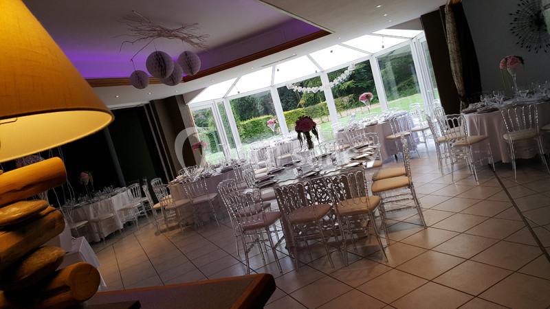 Salle de réception lumineuse avec tables dressées, chaises transparentes et décorations florales près de grandes baies…