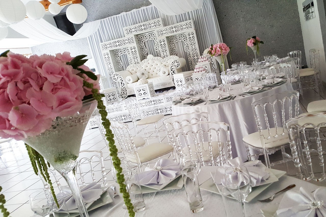 Salle de réception décorée avec des tables rondes, des chaises transparentes et des fleurs roses, devant une scène ornée.