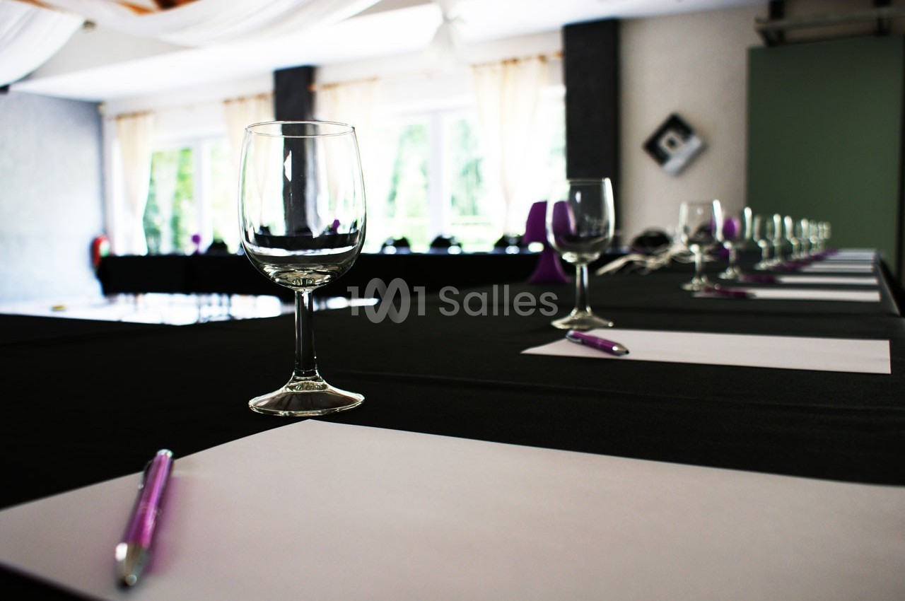 Salle de réunion avec des tables dressées, contenant des verres, des stylos et des feuilles blanches.