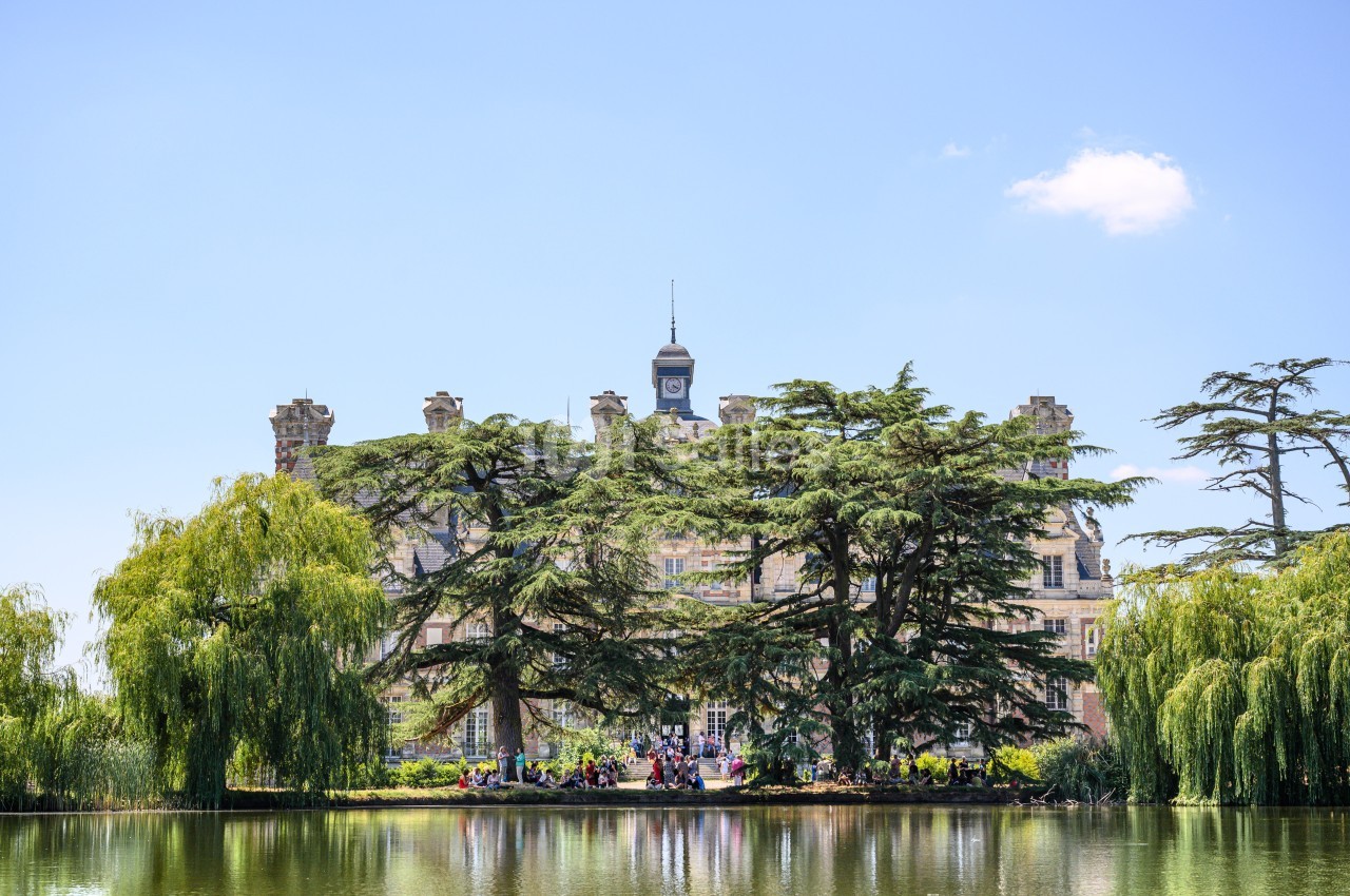 Façade d'un château entouré d'arbres, avec un plan d'eau au premier plan sous un ciel dégagé.