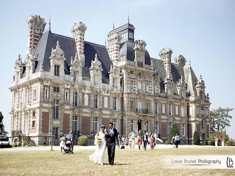 Un couple marche devant un château historique en pierre, entouré de visiteurs dans un parc verdoyant.