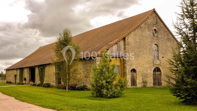 Grange en pierre avec toit en tuiles, entourée de pelouse, d'arbres et de buissons sous un ciel nuageux.