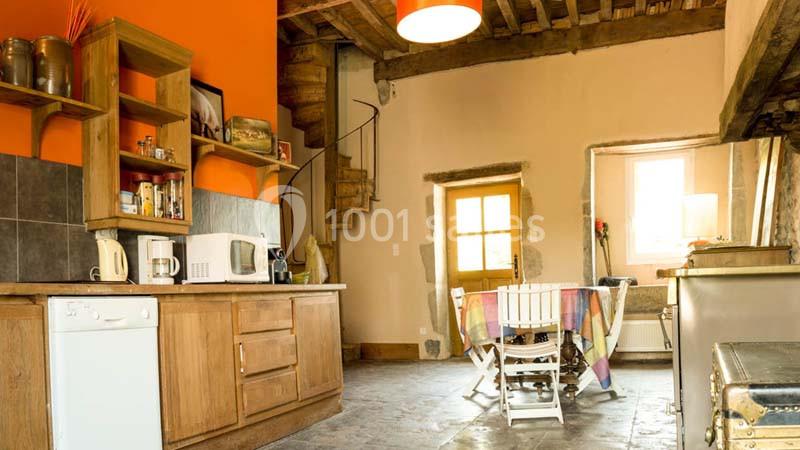 Cuisine rustique avec meubles en bois, table et chaises près d'une porte lumineuse, escalier en colimaçon visible au fond.