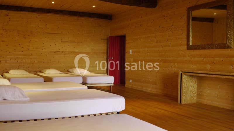 Salle en bois avec plusieurs lits simples alignés, éclairée par des spots au plafond, et un miroir doré au mur.