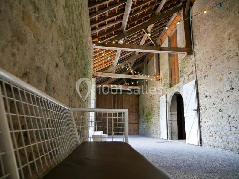 Intérieur d'une grange en pierre avec un banc en métal, des guirlandes lumineuses suspendues et des portes en bois ouvertes.